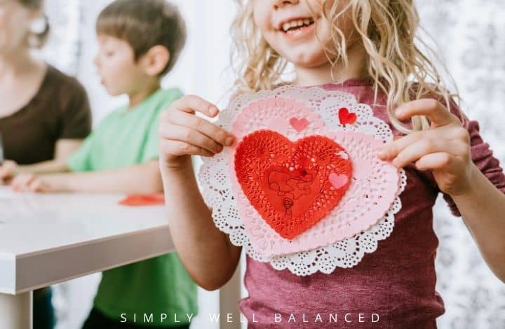 A little girl proudly shows off her Valentine's day craft - a pink and white heart and white doily. 