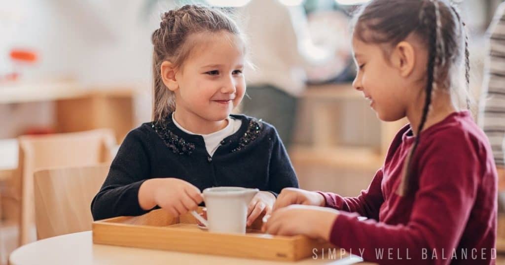 Two young girls engaged in open-ended, screen-free play with a small pitcher and tray at a wooden table.