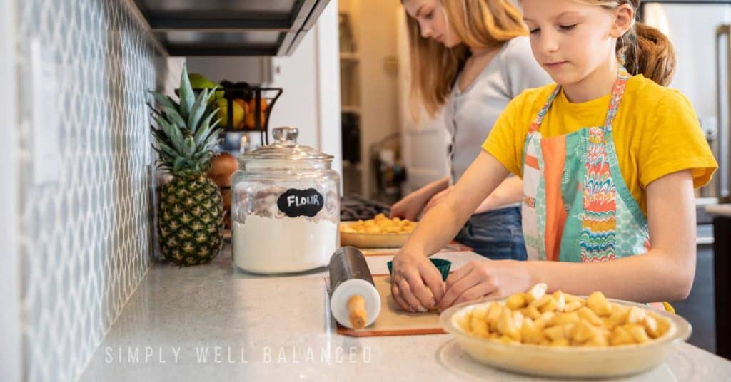 Two young girls making breakfast together in a bright kitchen with sliced apples on the counter.