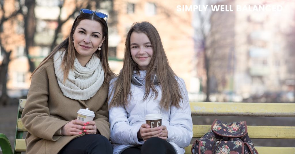Mother and teen daughter sitting on park bench in the fall holding warm drinks. Fall activities to do with your teen.