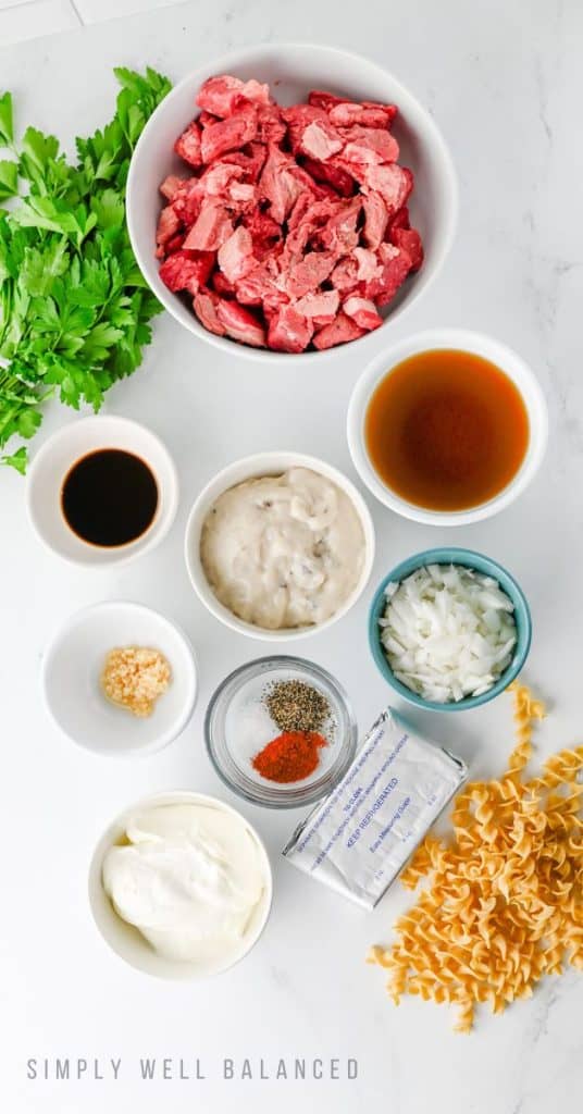 Flat lay image of the slow cooker beef stroganoff ingredients: stew meat, onion, garlic, cream of mushroom soup, beef broth, cream cheese and sour cream.